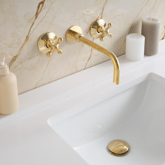 Timeless Brass Bathtub Faucet by Brasska Design with handcrafted unlacquered brass finish mounted on marble wall above white sink, with soap dispensers and candles.